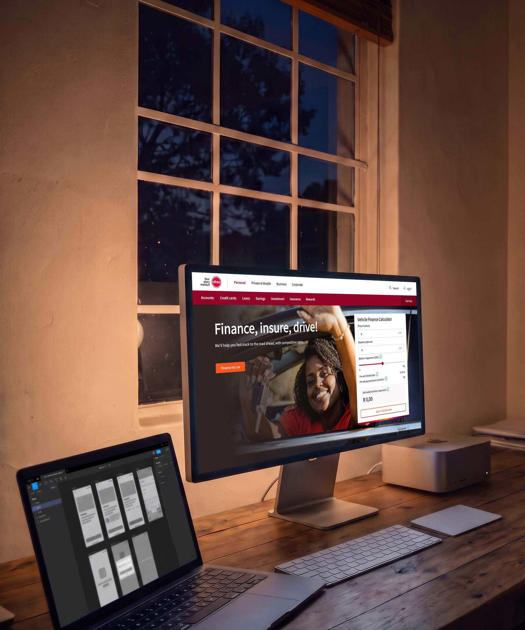 A designer’s desk at dusk lit only by its screens: a large display turned slightly away, showing Absa’s vehicle finance page headed Finance, insure, drive! with a vehicle finance calculator beside it, and a laptop at the left carrying app screens laid out in a design tool.