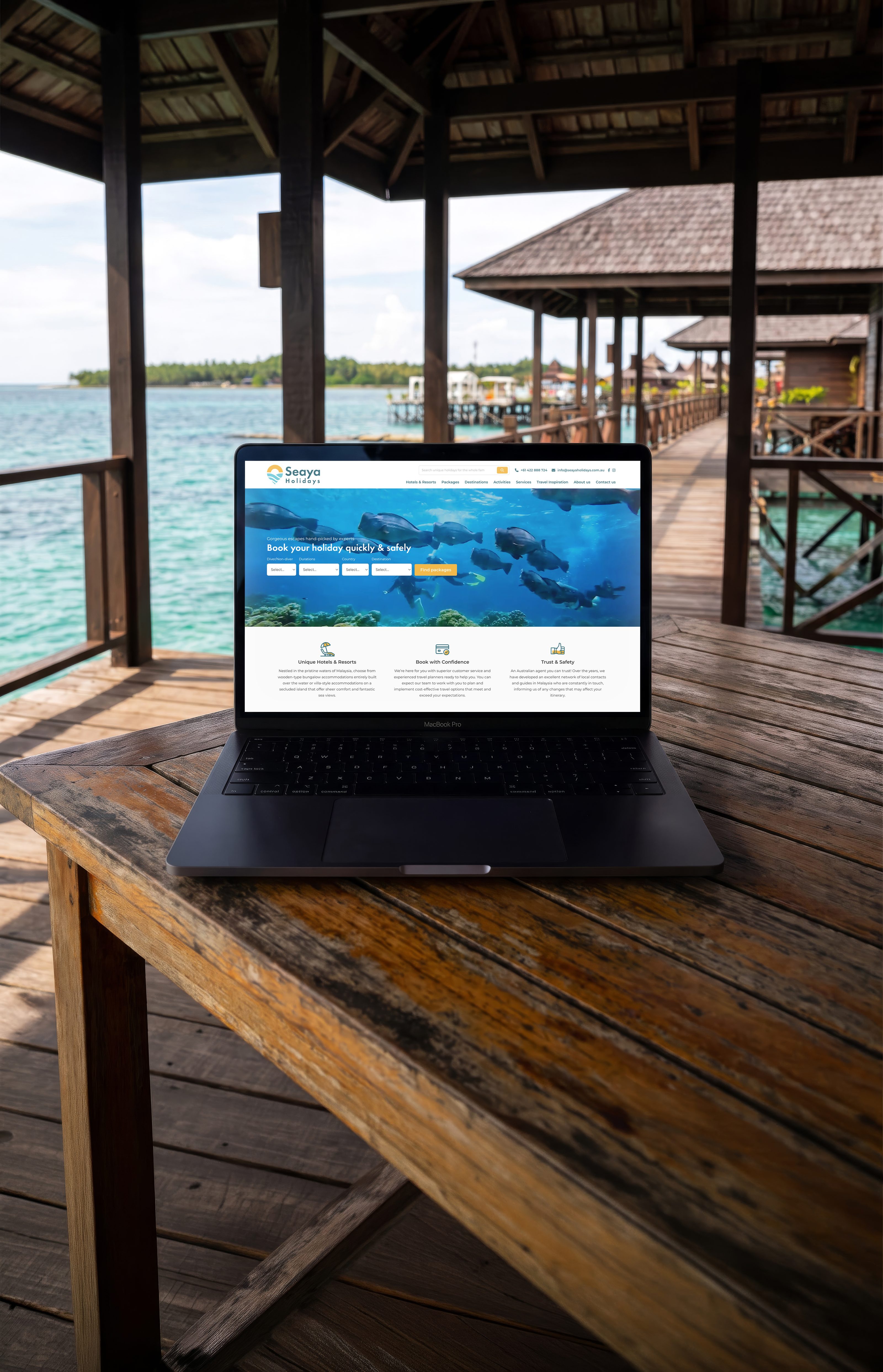 An open laptop on a weathered timber table in an overwater bungalow, its screen filled with the Seaya Holidays home page: the logo, the line Book your holiday quickly and safely over an underwater photograph of fish, a row of search fields beneath it and three blocks of reassurance below. Beyond the table, timber posts, a shingled bungalow on stilts and clear turquoise water.