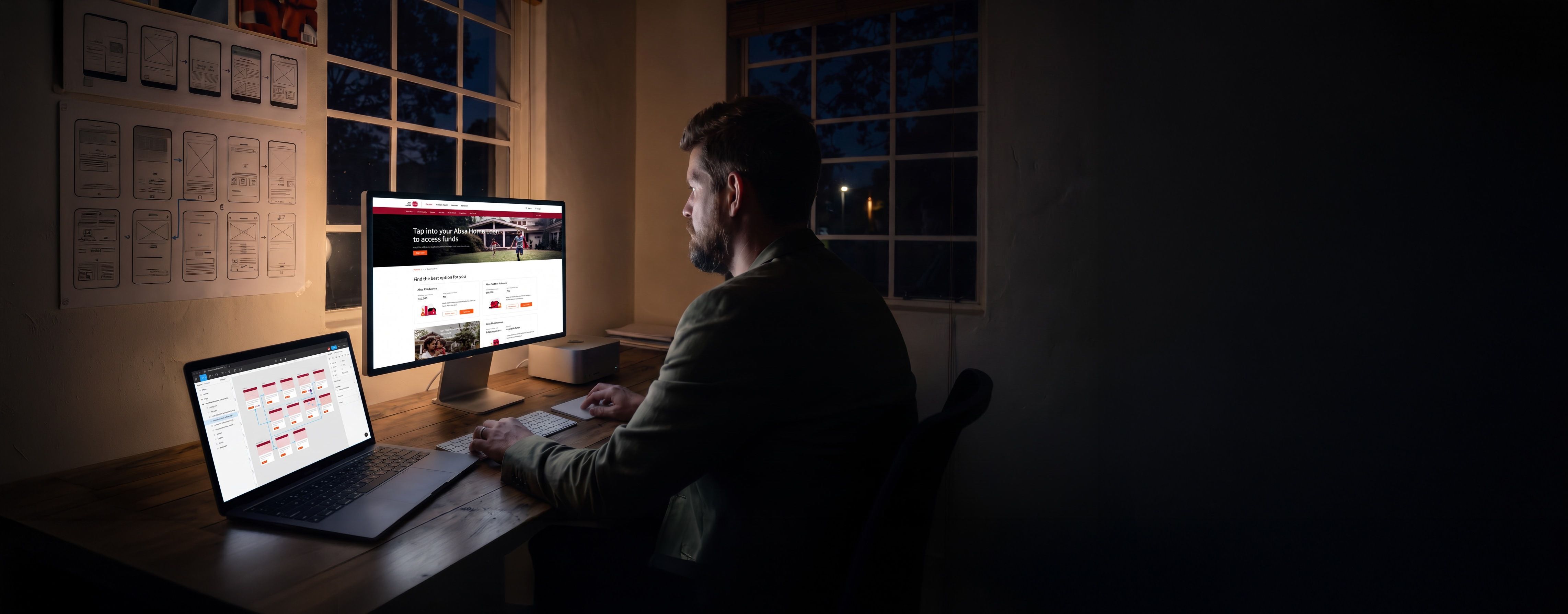 Will Derman at his desk in the studio at night, lit only by his screens: Absa's home loans page on the display, and on the laptop beside it the design file it was built in — dozens of small screens on a dark canvas, joined by connector lines into one long journey.