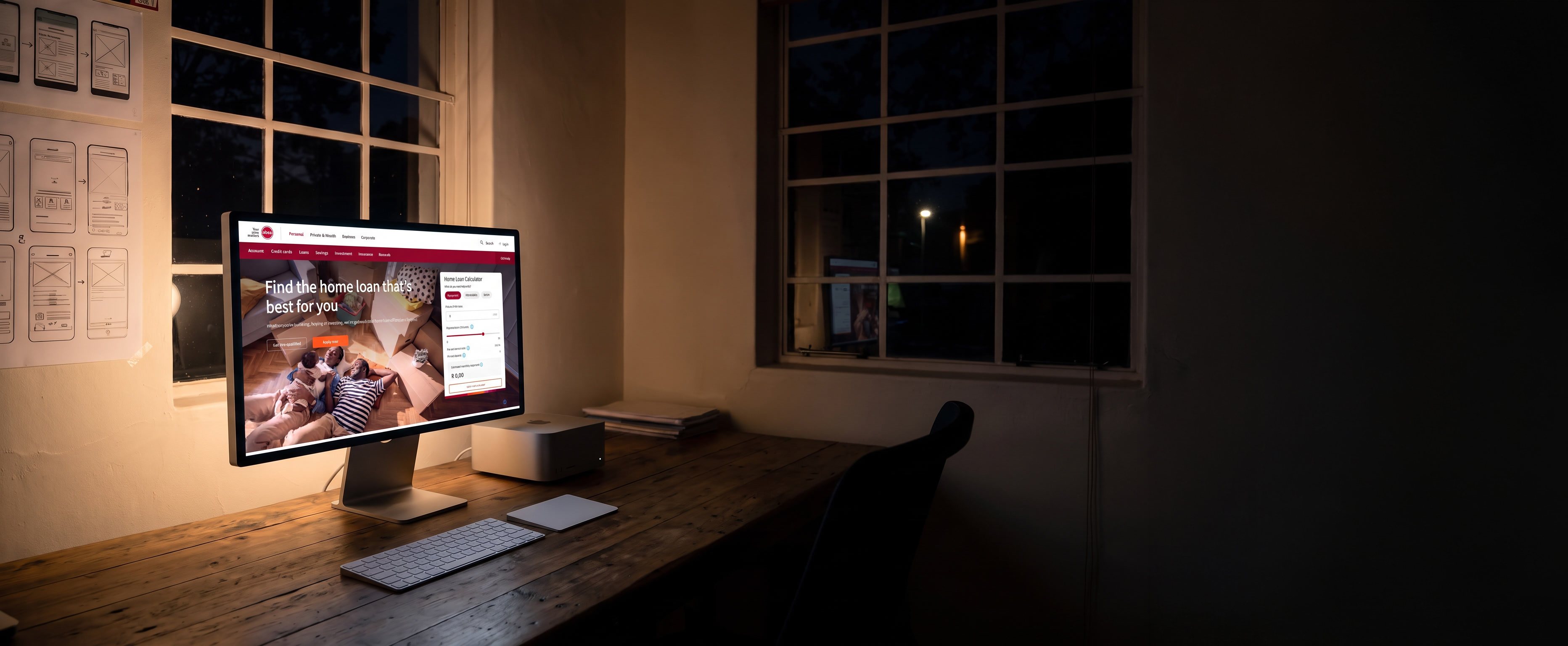 The studio desk at night, lit only by the screen: an Apple Studio Display and a Mac Studio on the timber desk, the Absa home loans page open on the display, and the wall of pinned wireframes beyond.